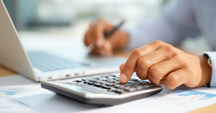 Photo of a man typing on a calculator with one hand and holding a pencil in his other hand while looking over a laptop