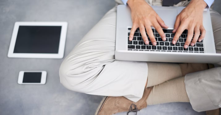 a corporate employee with more than one role at the company working on a laptop with an iPad and cell phone laying on the ground next to them as they sit cross-legged typing on their laptop
