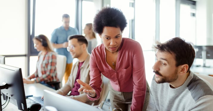 A female coworker talks with a male coworker at his desk in front of his computer at work 